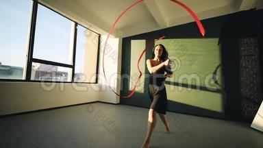 布鲁内特年轻女子舞蹈<strong>艺术体操</strong>，现代用红色丝带穿黑色连衣裙在窗户前。 太阳